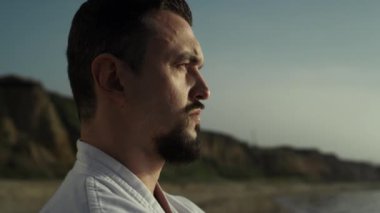 Dışarıda gün batımını seyreden atletik dövüşçülerin profili. Kumlu tepelerin önündeki vahşi adamın yüzünü kapatın. Düşünceli sporcu ayakta duran kumsaldaki eğitimden sonra doğanın tadını çıkarıyor..