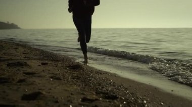 Unknown barefoot girl jogging on sandy beach. Fit woman legs running wearing leggings at sunset outside. Sporty female runner training on nature summer evening. Healthy cardio workout concept.