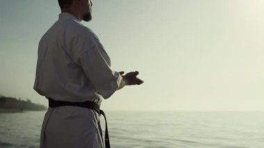 Karate dövüşçüsü güzel plajda gün batımının tadını çıkarıyor. Okyanus kenarında beyaz kimono giyen güçlü bir sporcu antrenmanı. Kumlu deniz kıyısında dövüş sanatları pratiği yapan atletik bir adam. Sağlıklı spor yaşam tarzı