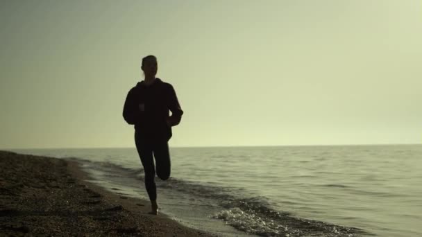 Kobieta z sylwetką na plaży o zachodzie słońca. Dziewczyna jogging wybrzeże letni wieczór. — Wideo stockowe