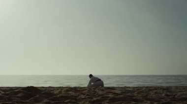 Karate man squat stretching legs on sandy beach. Athlete training flexibility.