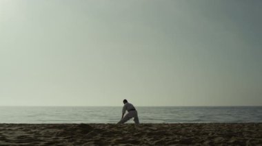 Sporty man doing warm-up squatting on sand. Karate fighter stretching on beach.
