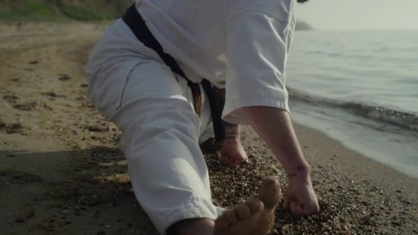 Flexibler, athletischer Mann, der sich aus nächster Nähe auf Sand dehnt. Karate-Kämpfer beim Training. — Stockvideo