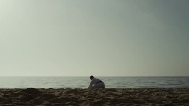 Karate man squat stretching legs on sandy beach. Athlete training flexibility. — Stockvideo
