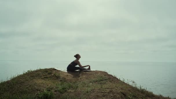 Kobieta ćwiczy jogę siedząc na zielonym wybrzeżu wzgórza. Sylwetka młoda dziewczyna rozciąganie — Wideo stockowe