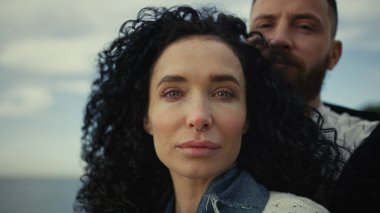 Deniz sahili arka planında bir çift kameraya bakıyor. Sevgililer aile gezisinde sarılırlar..