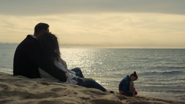 Aile deniz tatilinde gün batımını seyrediyor. İnsanlar kıyı şeridinde eğleniyor.