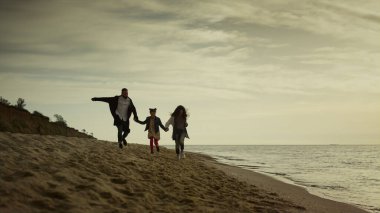 Aile gün batımında sahilde el ele tutuşur. Anne baba çocuk tatilde kum koşuyor..