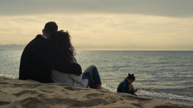 Rahat insanlar günbatımının tadını çıkarıyorlar. Kum kıyısında oturan bir aile..