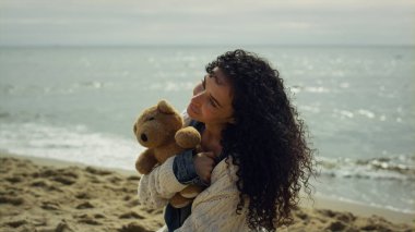 Latin kadın deniz sahilinde poz veriyor. Güzel bayan manken oyuncak ayıyı dışarıda tutuyor.