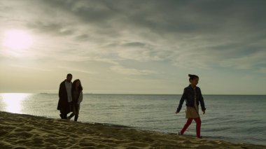 Neşeli bir aile sahil sahilinde yürüyor. Anne baba çocuğu doğa manzarasının tadını çıkarıyor.