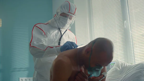 Lekarz zbadać19 pacjenta z powrotem stetoskop w masce ochronnej garnitur. — Zdjęcie stockowe