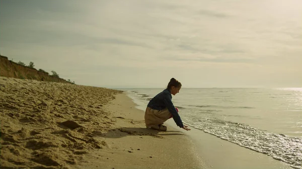 Dzieciak dotykający fal morskich na plaży o zachodzie słońca. Cute girl gry piasek na oceanie. — Zdjęcie stockowe