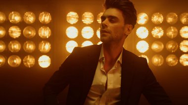 Seksi maço, sahne ışıkları altında düğmelerini açmış ceketini yakına çekiyor. Kameraya bakan seksi adam.