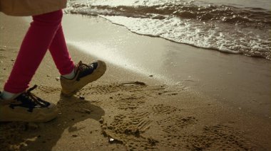 Deniz kumsalında yürüyen küçük kız bacakları. Çocuk ayakları uzaklaşıyor, dalgalar çarpıyor.
