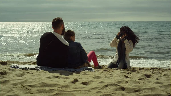 Young family taking pictures on sunny day beach. Mom dad child photographing sea