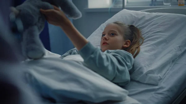 Хвора дівчина грає м'яку іграшку в клінічному ліжку. Дитина лежить в сучасній лікарняній кімнаті . — стокове фото