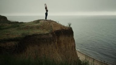 Athletic woman stand straightening up beach hill stretch hands to gray sky.