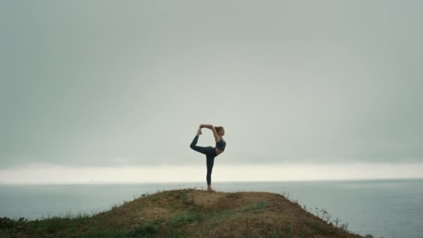Μια γυμνασμένη γυναίκα στέκεται στο ένα πόδι και τεντώνεται στο λόφο. Κορίτσι που κάνει γυμναστική. — Αρχείο Βίντεο