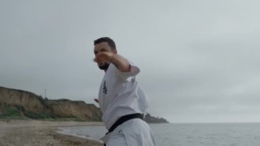Sportsman practicing hands punches on gloomy beach. Focused man workout karate.