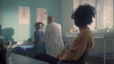Man doctor checking preteen kid reaction in office clinic. African mom waiting.