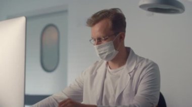 Masked doctor watching monitor in clinic close up. Man physician writing report 