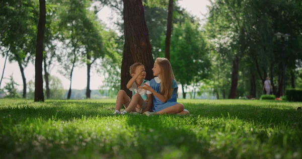 Skrattande barn sitter under träd i grön park. Glad aktiv barndom koncept. — Stockfoto