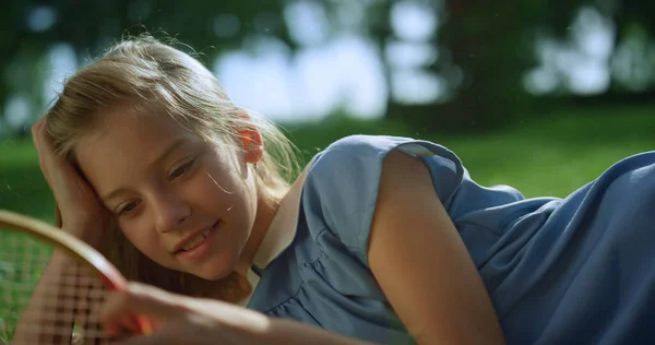Усміхнена дівчина лежить на зеленому газоні сама. Блондинка торкається мережі ракет крупним планом — стокове фото