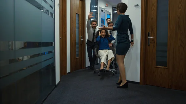 Multiethnic colleagues have fun ride office chair in corporate building hallway.