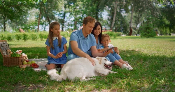 White dog lie down near family holding tablet on picnic. Father caress labrador. — Stock Photo, Image