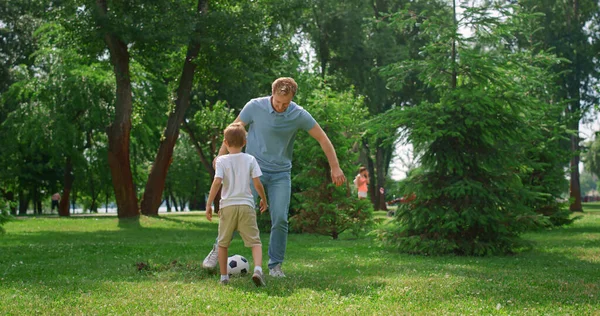 Glücklicher Vater, der seinem Sohn Fußball beibringt. Fröhlicher Junge gibt Ball an Papa auf Rasen weiter. — Stockfoto