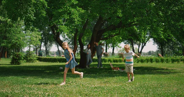 Aktiv flicka springer runt pojke på grönt gräs. Lekfulla barn ha kul på picknick. — Stockfoto