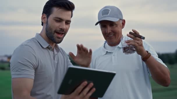 Leende män ringer online video innehav tablett på golfbanan. Förmögen idé. — Stockvideo