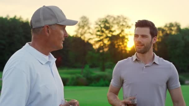 Lachende golfgroep aan het praten op Sunset Fairway. Twee spelers genieten van een drankje op koers. — Stockvideo