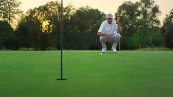 Oude golfsporter die een spelletje speelt op de groene baan. Golfer genieten van de zomer zonsondergang. — Stockvideo