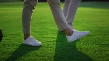 Yeşil çimlerin üzerinde yürüyen iki golfçü bacağı. Golf takımı golf sahasında spor malzemeleri taşıyor. Spor giyim üniforması giyen kimliği belirsiz adamlar. Lüks yaşam tarzı hobi eğlence konsepti.