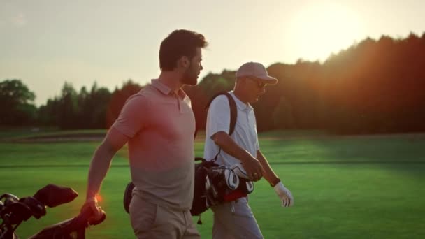 商人走在外面的高尔夫球场上 两名职业选手穿着运动服带着俱乐部 高尔夫球队的朋友们在夏日落日场讨论丰富的生活方式爱好 体育伙伴关系概念 — 图库视频影像