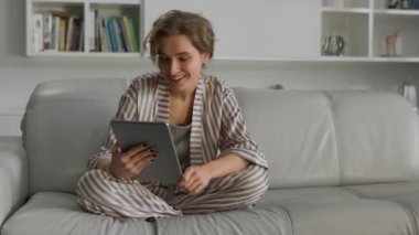 Ev hanımı, oturma odasında elinde tablet bilgisayarla sohbet etmekten hoşlanıyor. Gülümseyen çekici kadın evinde video görüşmesi yaparak dinleniyor. Neşeli kız öğrenci sabahları pijama giyer..