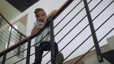 Merdivenlerden eğilmiş okul koridoruna bakan sevimli bir öğrenci. Meraklı çocuk koridorda tek başına dikiliyor. İlgili genç öğrenci molada sıkıldı. Modern çocukluk kavramı.