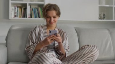 Yakından bir kadın cep telefonundan mesaj yazıyor. Gülümseyen kız oturma odasında online sohbetten hoşlanır. Neşeli çekici hipster rahat evinde dinlenirken iyi haberler okuyor. Akıllı telefon kullanan mutlu kadın. Kanepede rahatla..