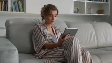 Oturma odasında dijital tablet bilgisayarda banka hesabını kontrol eden sürpriz kız. Tablet bilgisayar kullanan çekici bir kadın pijamalı sosyal medyayı kontrol ediyor. Genç yetişkinler hafta sonları dinlenme evlerinde eğlenirler.