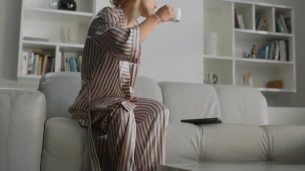 女性はコーヒーカップで家で働き始める 居心地の良い遠隔地の職場だけでマネージャー パジャマを着た美しいリラックスした女性がコーヒーを飲みながらノートパソコンを開きます 可愛いです女の子でベッドウェアでソファに座って — ストック動画