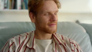 Mutlu hipster dinlenme koltuğu evde yakın çekim. Gülen kızıl adam kanepeye uzan. Neşeli sakallı serbest çalışan uzak bir iş yerinde işten sonra dinleniyor. Tatmin olmuş genç baba, sakin atmosferin tadını çıkar.