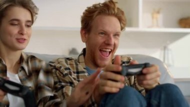 Oyuncu çift evde yakın çekimde joystickçilik oynamaktan hoşlanıyor. Heyecanlı oyuncular eğleniyor. Güzel rekabetçi eş mutlu kızıl koca online nişancı aldatması oynuyor. Oyuncular turnuvası hafta sonu akşamı.