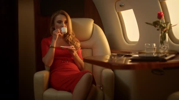 黄金の太陽の下でファーストクラスの旅をお楽しみください プライベート飛行機でコーヒーを飲む赤いドレスの豊かな女性 成功した実業家は電子メールのソーシャルメディアをリラックスさせます 裕福なブロンド使用スマートフォン — ストック動画