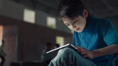 Oyun oynayan heyecanlı genç çocuk okul salonunda elinde tablet tutuyor. Dijital cihaz kullanarak tek başına bankta oturan neşeli, takıntılı öğrenci. İlkokul öğrencisi, modern aletlerle video izliyor..