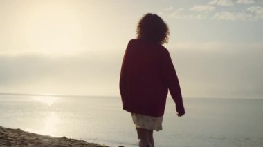 Gündoğumunda deniz kıyısında yürüyen sıradan bir kız. Pozitif kadın el kaldırıyor. Başarıyı kutlayan kadın. Genç bayan özgürlüğün tadını çıkarıyor. Güneş ışıkları okyanus yüzeyine düşüyor. Zafer konsepti