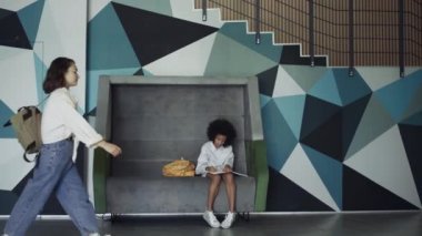 Düşünceli Afrikalı Amerikalı öğrenci modern okul koridorunda bankta oturuyor. Kıvırcık liseli kız koridorda ödev yazıyor. Yaratıcı odaklı genç kız ara verildiğinde deftere resim çiziyor..