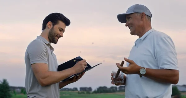 Two men talking golf course outside. Golfers friends analyze game result at sunset field close up. Sport team interviewing taking notes on country club landscape view. Leisure relax activity concept.