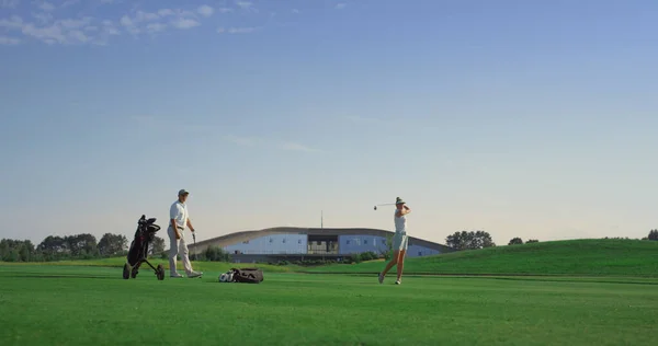 Two golf players training at course field. Couple playing sport game in sunset. Golfers group swinging hit golfing ball on fairway nature. Rich people practicing activity outdoors. Leisure fun concept